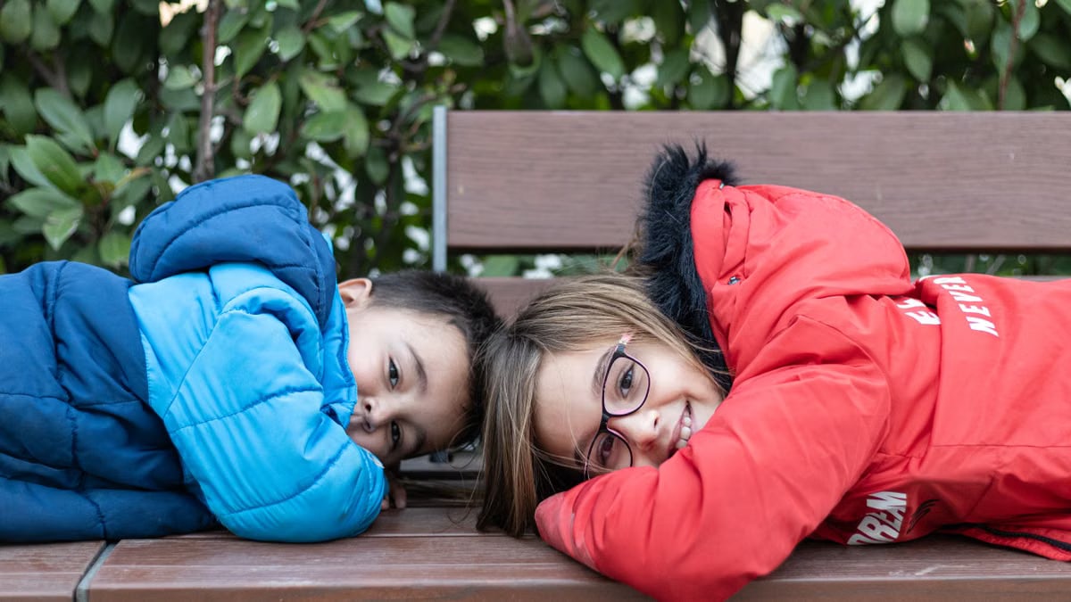 Two kids in colorful jackets lying on a wooden bench outdoors beside leafy greenery.